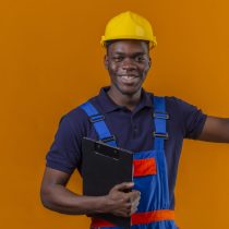 young african american builder man wearing construction uniform and safety helmet standing with clipboard raising hand clenching fist smiling standing with happy face celebrating victory win